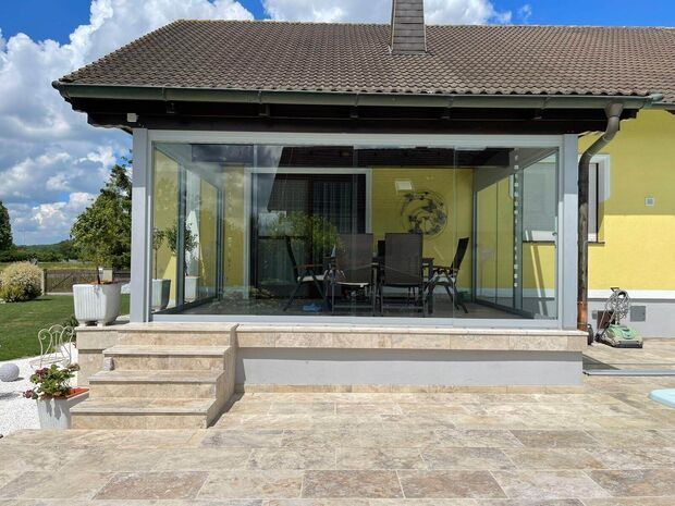 Eine Terrasse mit Glastüren und einem Tisch, umgeben von Pflanzen und Sonnenlicht.